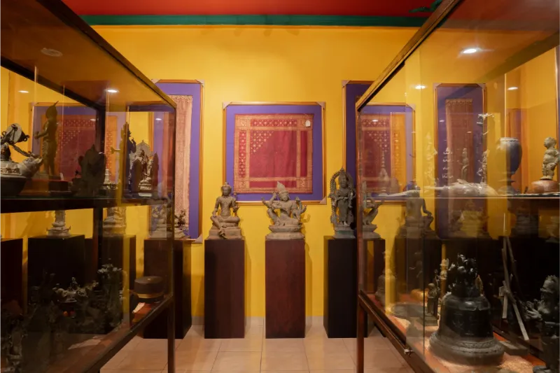 Ancient Hindu-Buddhist bronze statues on dark pedestals with framed traditional textiles, Museum Peter Bloch Collection.