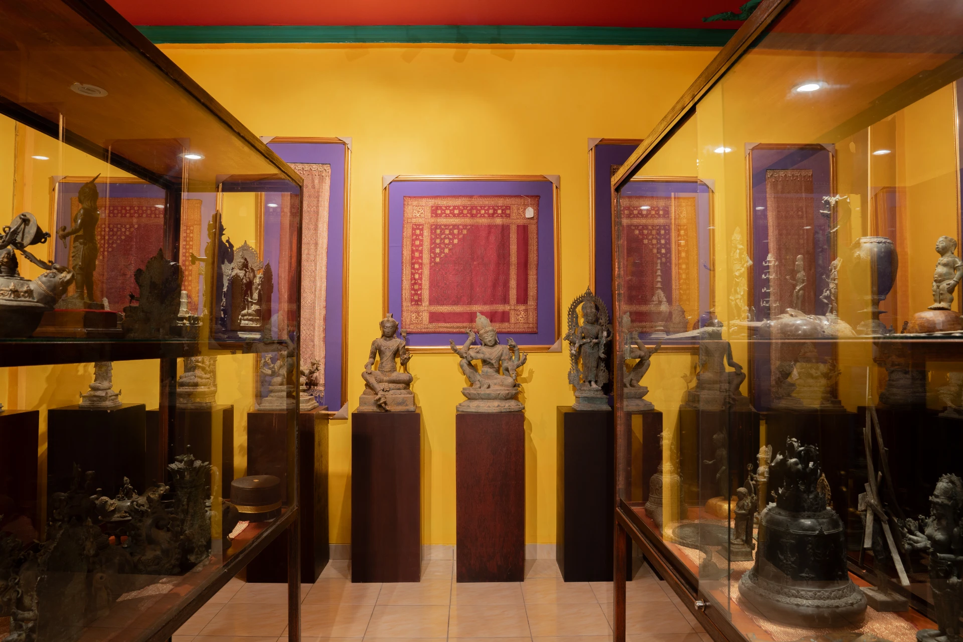Ancient Hindu-Buddhist bronze statues on dark pedestals with framed traditional textiles, Museum Peter Bloch Collection.